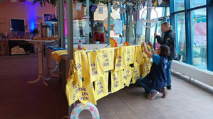 Zwei Personen dekorieren einen mit gelben T-Shirts bedeckten Tisch mit der Aufschrift "THE UNSEEN" in einem hellen Raum mit großen Fenstern. Eine Person hängt die Dekoration an einem runden Rahmen auf, während eine andere die Gegenstände in der Nähe des Tisches zurechtrückt.