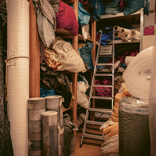 Ein Lagerraum voller Stoffrollen und verschiedener Textilien, die auf Regalen und dem Boden gestapelt sind. In der Mitte steht eine Leiter, um an die höheren Regale zu gelangen. Der Raum ist vollgestopft mit Materialien in verschiedenen Farben und Texturen.