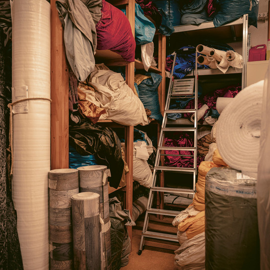 Ein Lagerraum voller Stoffrollen und verschiedener Textilien, die auf Regalen und dem Boden gestapelt sind. In der Mitte steht eine Leiter, um an die höheren Regale zu gelangen. Der Raum ist vollgestopft mit Materialien in verschiedenen Farben und Texturen.