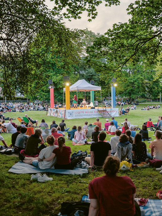 Eine große Menschenmenge hat sich draußen auf einem grasbewachsenen Hügel versammelt, sitzt auf Decken und Stühlen und sieht sich eine Aufführung auf einer kleinen, von Bäumen umgebenen Bühne an. Es herrscht eine lebhafte Stimmung, und die Menschen genießen die Open-Air-Veranstaltung an einem hellen Tag.