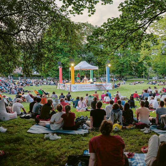 A large group of people sits on the grass in a park, watching a performance on a small, elevated stage surrounded by trees. The audience is spread out on blankets, enjoying the outdoor event under a clear sky.