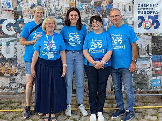 Five people stand smiling in front of a mural, all wearing blue "Chemnitz 2025 VOLUNTEER" shirts advertising Chemnitz as European Capital of Culture 2025.