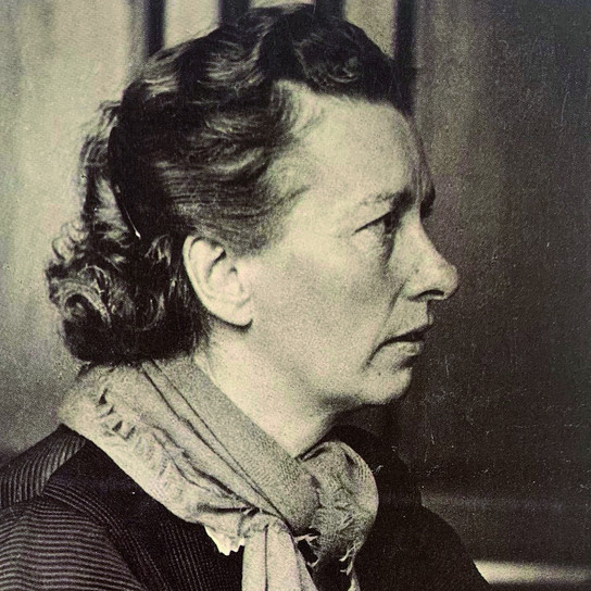 Ein Schwarz-Weiß-Porträt einer Frau mittleren Alters im Profil. Sie hat welliges, nach hinten frisiertes Haar und trägt ein Tuch um den Hals. Der Hintergrund ist leicht verschwommen, was ihren nachdenklichen Gesichtsausdruck unterstreicht.