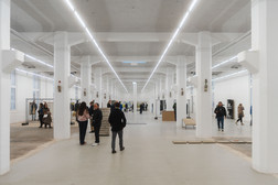 A spacious, well-lit industrial hall with white pillars and high ceilings, where people are scattered around, some chatting, others taking a walk or looking at the exhibitions on the walls.
