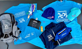 Eine Sammlung von blauen "Chemnitz 2025 Volunteer"-Shirts, ein Rucksack, ein Regenponcho, eine blaue Mütze, eine türkisfarbene Kappe, eine marinefarbene Jacke, eine Gürteltasche, ein Regenschirm und Sonnencreme, alles auf einem Tisch ausgebreitet.
