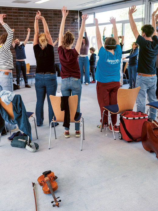A group of people in a classroom stand in a circle with their arms raised. Chairs are arranged around them, and musical instruments, including a violin and cello, are on the floor. The room is well-lit with large windows.