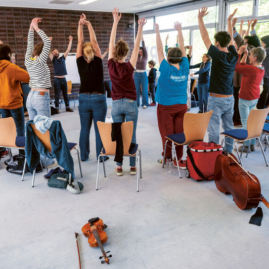 A group of people in a classroom stand in a circle with their arms raised. Chairs are arranged around them, and musical instruments, including a violin and cello, are on the floor. The room is well-lit with large windows.
