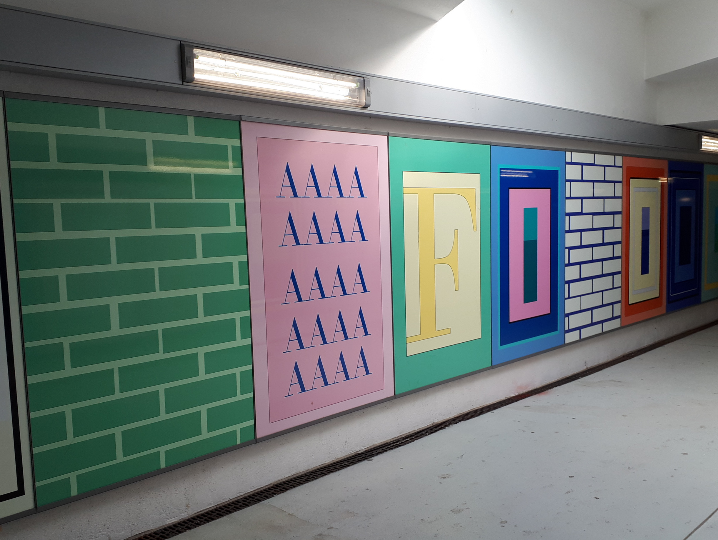 Eine Flurwand ist mit bunten Paneelen dekoriert, darunter grün-weiße Ziegelmuster, wiederholte „AAAA“-Buchstaben in Pink und große stilisierte Buchstaben mit der Aufschrift „FOOD“ in Gelb, Rot, Blau und Orange.