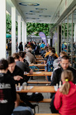 People sit in rows at long tables outside and play chess under a covered walkway. The square is bustling with players and spectators, and trees can be seen in the background.