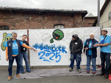 Fünf Personen stehen vor einem Graffiti-Wandbild mit der Aufschrift "Volunteer 2025" auf einer weißen Plane; drei zeigen auf das Wandbild, alle tragen blaue Freiwilligenhemden oder -abzeichen, und die Szene spielt sich im Freien zwischen Backsteinbauten ab.