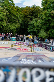A skateboarder performs a trick over a rail in a graffiti-covered skate park while a crowd watches and trees can be seen in the background.