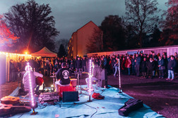 Eine Band spielt in der Abenddämmerung auf einer Freilichtbühne, umgeben von Lichterketten, und eine große Menschenmenge schaut zu. Im Hintergrund sind Bäume, Gebäude und ein Zelt zu sehen.
