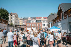 Eine Gruppe von Menschen versammelt sich an einem sonnigen Tag im Freien in einem städtischen Gebiet. Im Hintergrund sind bunte alte Gebäude und ein blaues Zelt zu sehen. Die Menge scheint an einer Veranstaltung oder einer Tournee teilzunehmen.