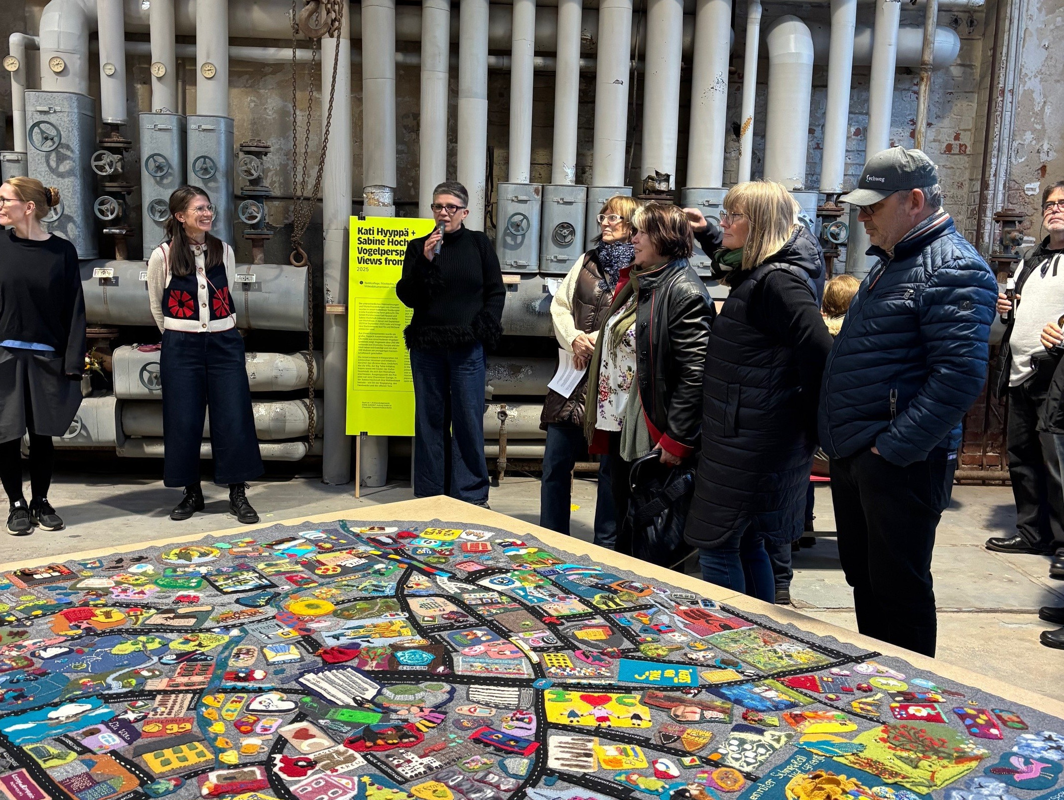 Eine Gruppe von Personen steht in einem Industrieraum mit freiliegenden Rohren und betrachtet ein bunt besticktes Textilkunstwerk, das auf einem Tisch im Vordergrund ausgestellt ist. Im Hintergrund ist ein leuchtend gelbes Schild zu sehen.