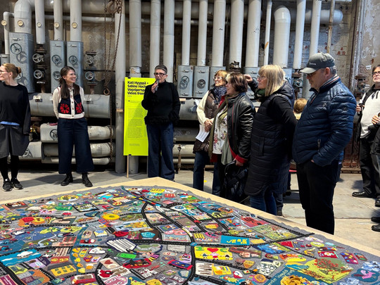 Eine Gruppe von Personen steht in einem Industrieraum mit freiliegenden Rohren und betrachtet ein bunt besticktes Textilkunstwerk, das auf einem Tisch im Vordergrund ausgestellt ist. Im Hintergrund ist ein leuchtend gelbes Schild zu sehen.