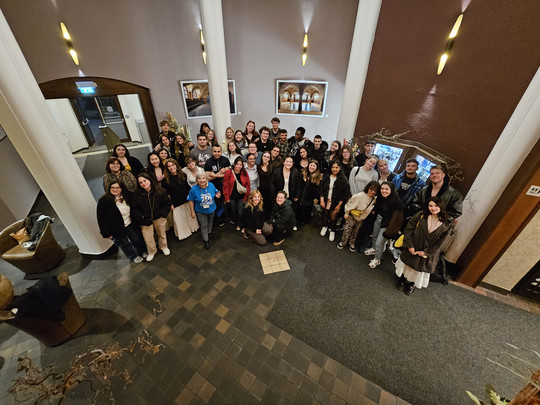 Eine große Gruppe von Menschen posiert für ein Foto in der Lobby eines Gebäudes mit gefliesten Böden, weißen Säulen und Wanddekoration. Die meisten lächeln und blicken in die Kamera, einige stehen, andere sitzen vorne.