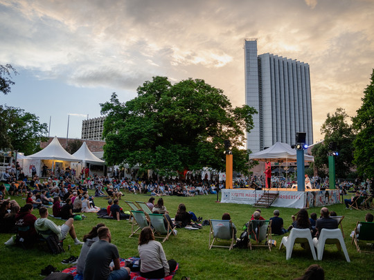 Eine große Menschenmenge sitzt im Gras und auf Stühlen bei einer Veranstaltung im Freien und beobachtet eine Bühnenvorführung bei Sonnenuntergang. Im Hintergrund sind Bäume, Zelte und ein hohes modernes Gebäude zu sehen.