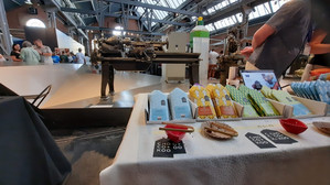 Ein Schautisch mit verpackten Schokoladentafeln und Kostproben in einer industriellen Markthalle mit Menschen und alten Maschinen im Hintergrund.
