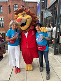 Vor einem Backsteingebäude mit Glaseingang stehen zwei Frauen in blauen Freiwilligenhemden neben einer Person in einem Löwenmaskottchenkostüm, alle lächeln und halten Veranstaltungsbroschüren in der Hand.