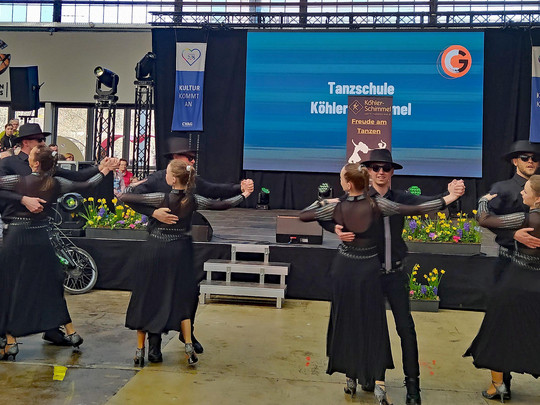 Vier Paare in schwarzen Outfits führen auf einer Indoor-Bühne einen Gesellschaftstanz auf. Zuschauer schauen zu, auf einer Leinwand dahinter ist „Tanzschule Köhler-Schimmel“ zu sehen. Der Saal ist mit Blumen und Bannern geschmückt.