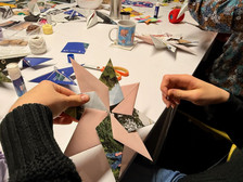 Zwei Personen falten und fügen bunte Papiersterne an einem Tisch zusammen, der mit Bastelmaterial gefüllt ist, darunter Scheren, Klebstoff, ein Becher und verstreute Papierstücke.