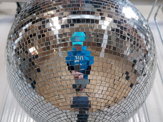 Eine Person in blauem Hemd und Hut spiegelt sich in einer großen, glänzenden Discokugel aus verspiegelten Kacheln, im Hintergrund sind ein Holzboden und Rohre zu sehen.