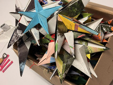 Eine Pappschachtel ist mit bunten, dreidimensionalen Origami-Sternen aus verschiedenen gemusterten und festen Papieren gefüllt. Schere, Klebeband und Bastelwerkzeug liegen auf dem Tisch daneben.