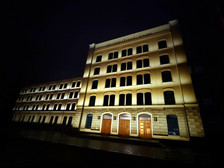Ein großes, mehrstöckiges Gebäude, das nachts von warmen Lichtern beleuchtet wird, mit Rundbogenfenstern und drei großen Holztüren. In der Nähe des Daches ist das Schild "UNIWERSYTECKA BIBLIOTEKA" zu sehen. Die Umgebung ist dunkel.