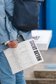 A person dressed in light blue is holding a folded piece of paper with the title "SICHT BETON" printed in bold and is carrying a dark blue bag over his shoulder.