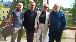 Vier Erwachsene stehen an einem sonnigen Tag Seite an Seite im Freien und lächeln in die Kamera. Im Hintergrund sind Bäume, eine Wiese und gelbe Gebäude zu sehen. Sie tragen legere und halboffizielle Kleidung.