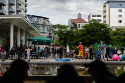 At an outdoor civic event, a crowd gathers by a pond to watch a street performer blow bubbles. People of all ages enjoy the lively scene surrounded by modern buildings and green spaces.