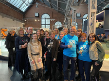 Eine Gruppe von Menschen posiert gemeinsam in einem Innenraum bei einer Veranstaltung. Einige tragen blaue "Chemnitz 2025 Volunteer"-T-Shirts, während andere Freizeit- oder Geschäftskleidung tragen. Sie lächeln und stehen in einem gut beleuchteten Ausstellungsraum.