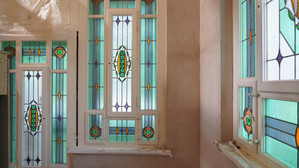 Sunlight shines through decorative stained glass windows with geometric and floral patterns in shades of aquamarine, green, orange and purple, illuminating an interior with beige-coloured walls.