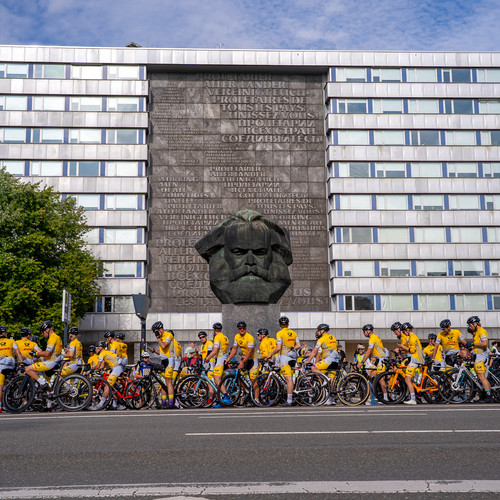 Eine Gruppe von Radfahrern in gelben Trikots versammelt sich mit ihren Rädern vor einem großen Karl-Marx-Denkmal, das in ein modernes Gebäude mit einem Gitter aus Fenstern eingebettet ist. Links sind Bäume unter blauem Himmel zu sehen.