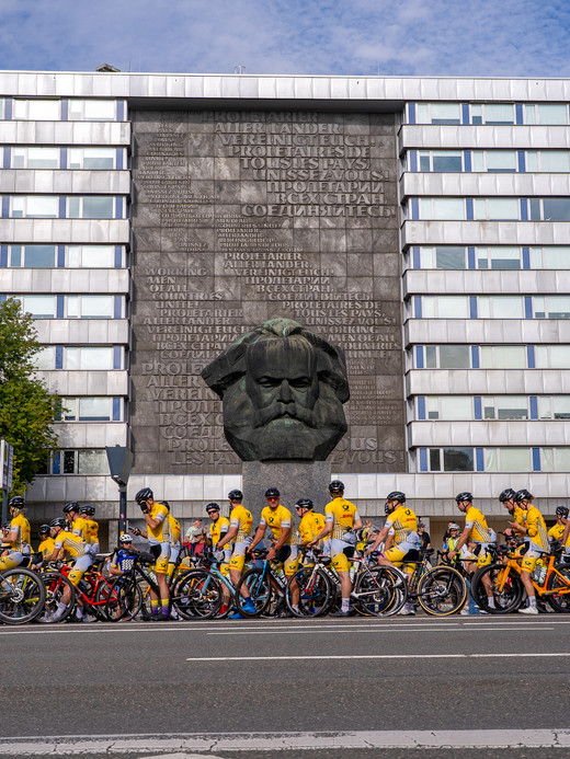 Eine Gruppe von Radfahrern in gelben Trikots versammelt sich mit ihren Rädern vor einem großen Karl-Marx-Denkmal, das in ein modernes Gebäude mit einem Gitter aus Fenstern eingebettet ist. Links sind Bäume unter blauem Himmel zu sehen.