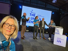 Eine Frau macht ein Selfie bei einer Indoor-Veranstaltung. Im Hintergrund stehen vier Personen auf einer Bühne vor einer großen Leinwand, auf der "The Unseen Chemnitz Kulturhauptstadt Europas 2025" zu sehen ist. Die Umgebung wirkt festlich und professionell.