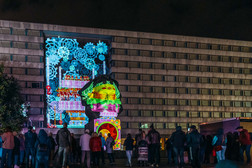 Eine Gruppe von Menschen steht in der Nacht und betrachtet bunte, animierte Lichtprojektionen von Zahnrädern und abstrakten Mustern, die an der Seite eines großen, modernen Gebäudes angebracht sind.