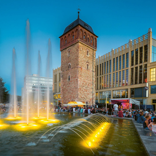 A historic tower stands beside a modern building at dusk. A vibrant fountain with illuminated jets fills the foreground. A large crowd gathers near an outdoor stage, enjoying a lively evening atmosphere.