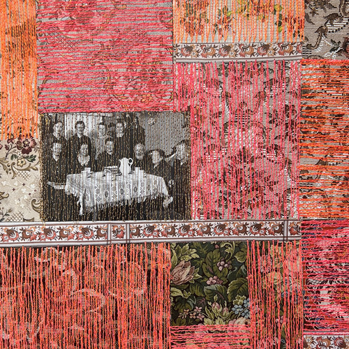 Ein Patchwork aus floralen und gemusterten Stoffquadraten, hauptsächlich in Rosa- und Orangetönen, mit vertikal herabhängenden orangefarbenen Fäden. Ein Quadrat enthält ein Schwarzweißfoto von Personen, die an einem Tisch sitzen.