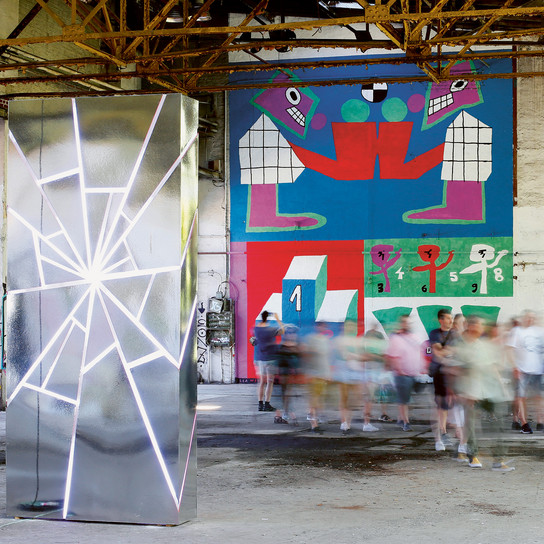 A large metallic sculpture with neon lines stands in an industrial space. Bright, abstract murals cover the walls. Blurry figures move nearby, creating a sense of motion. The atmosphere is artistic and vibrant.