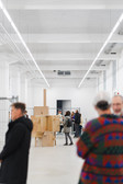 A group of people are standing and chatting in a bright, modern art gallery with high ceilings. Cardboard boxes and sculptures are displayed in the background. The scene has a casual and informal feel.