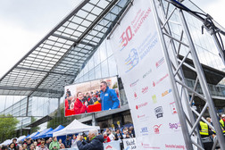 A large outdoor marathon event with a crowd of people, sponsor banners and a large screen on which two people are speaking. The scene takes place on a cloudy day in front of a modern glass building.