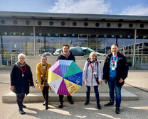Fünf Personen stehen im Freien vor einem modernen Gebäude und lächeln in die Kamera. Eine Person hält einen bunten Regenschirm. Sie alle tragen Schlüsselbänder und Mäntel, und hinter ihnen ist eine große Skulptur zu sehen.