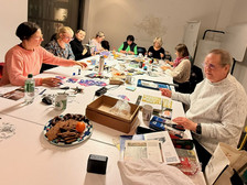 Eine Gruppe von Menschen sitzt in einem gut beleuchteten Raum um Tische mit Bastelmaterial, Snacks und Getränken und ist mit kreativen Tätigkeiten beschäftigt. Einige schneiden Papier aus, andere zeichnen oder unterhalten sich.