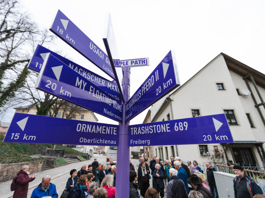 Eine Gruppe von Menschen steht um einen violetten Wegweiser am PURPLE PATH in Hainichen mit mehreren Pfeilen, die in verschiedene Richtungen weisen und jeweils mit Zielnamen und Entfernungen in Kilometern beschriftet sind.