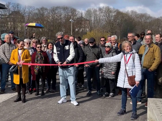 Eine Gruppe lächelnder Menschen, während ein Mann und eine Frau im Freien ein rotes Band halten und durchschneiden, um die Eröffnung eines Gehwegs oder einer Veranstaltung zu markieren. Im Hintergrund sind Bäume und Gebäude zu sehen.