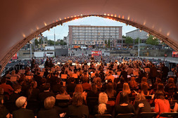 Blick von hinten auf ein Orchester, das auf einer Freilichtbühne unter einem weißen Vordach spielt, mit Blick auf ein großes, sitzendes Publikum in einem städtischen Gebiet, in dem im Hintergrund Gebäude, Bäume und Straßenlaternen zu sehen sind.