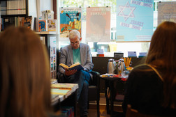 An older man with grey hair and glasses sits in a bookshop and reads a book to an audience. Around him are bookshelves, posters and a table with stationery. The photo is taken from behind two listeners.