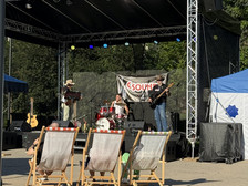 Drei Musiker treten auf einer Außenbühne unter einem schwarzen Baldachin auf, mit Schlagzeug, Gitarre und Bass. Hinter ihnen hängt ein "Sound"-Banner. Mehrere Leute schauen von Liegestühlen im sonnigen Zuschauerbereich aus zu.
