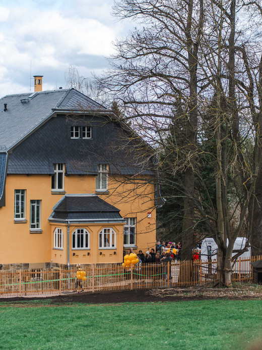 Ein großes gelbes Haus mit dunklem Dach ist von kahlen Bäumen und einer Wiese umgeben. Gelbe Luftballons schmücken den umzäunten Hof, und mehrere Menschen haben sich in der Nähe des Eingangs versammelt. Der Himmel ist teilweise bewölkt.
